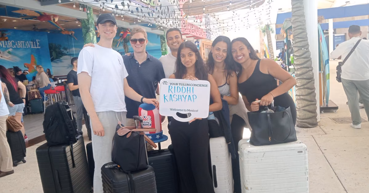 Six smiling people with suitcases at an airport, one holding a “Riddhi Kashyap, Welcome to Mexico” sign; tropical decor behind.