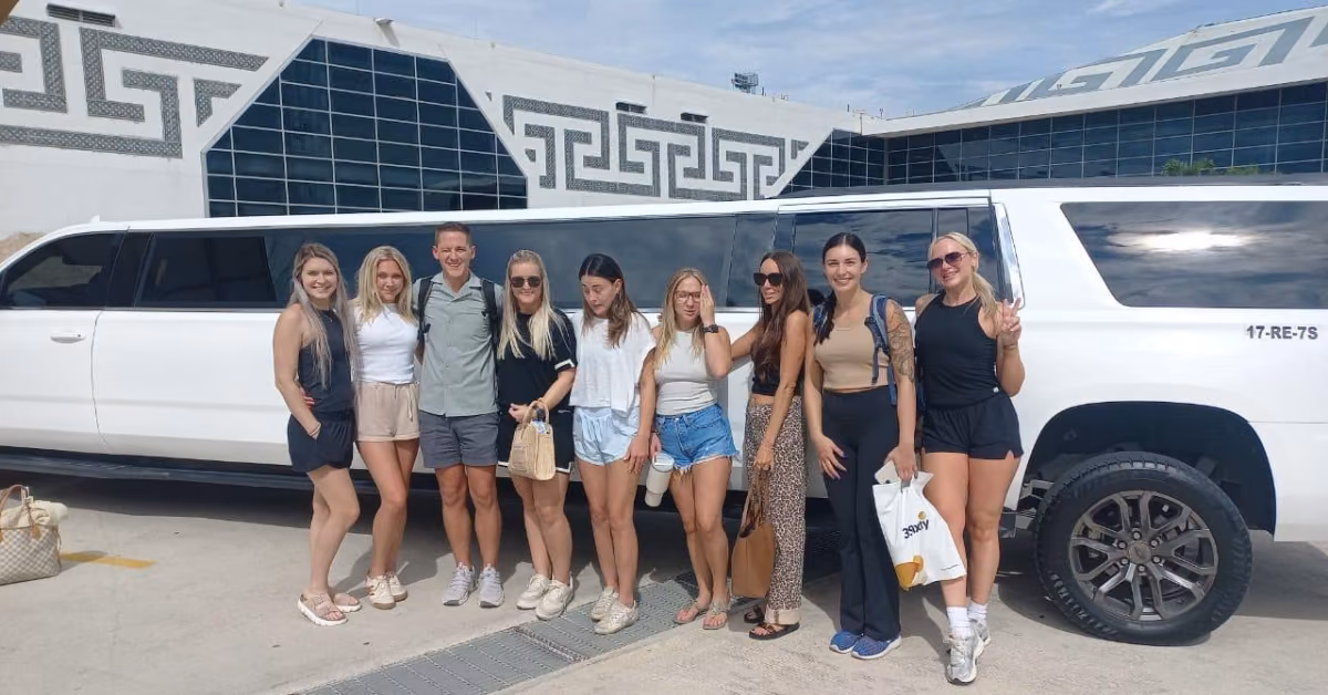 Eight people smile in front of a white limousine by a modern building. Most are casually dressed and holding bags under clear skies.