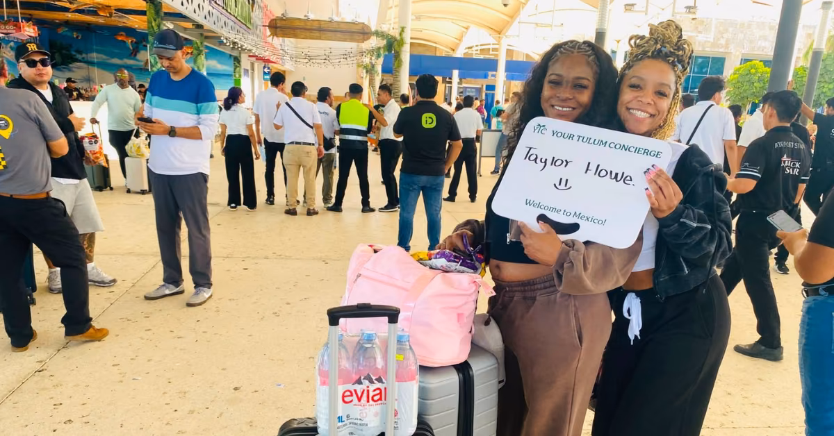 Two women smile with a Welcome to Mexico sign at an airport, standing by luggage and water as travelers pass by in the background.