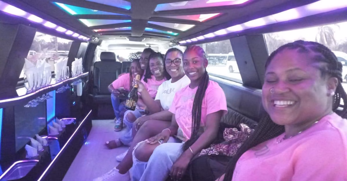 Five women in matching pink shirts smile together in a bright limousine with neon lights and champagne flutes along the side.