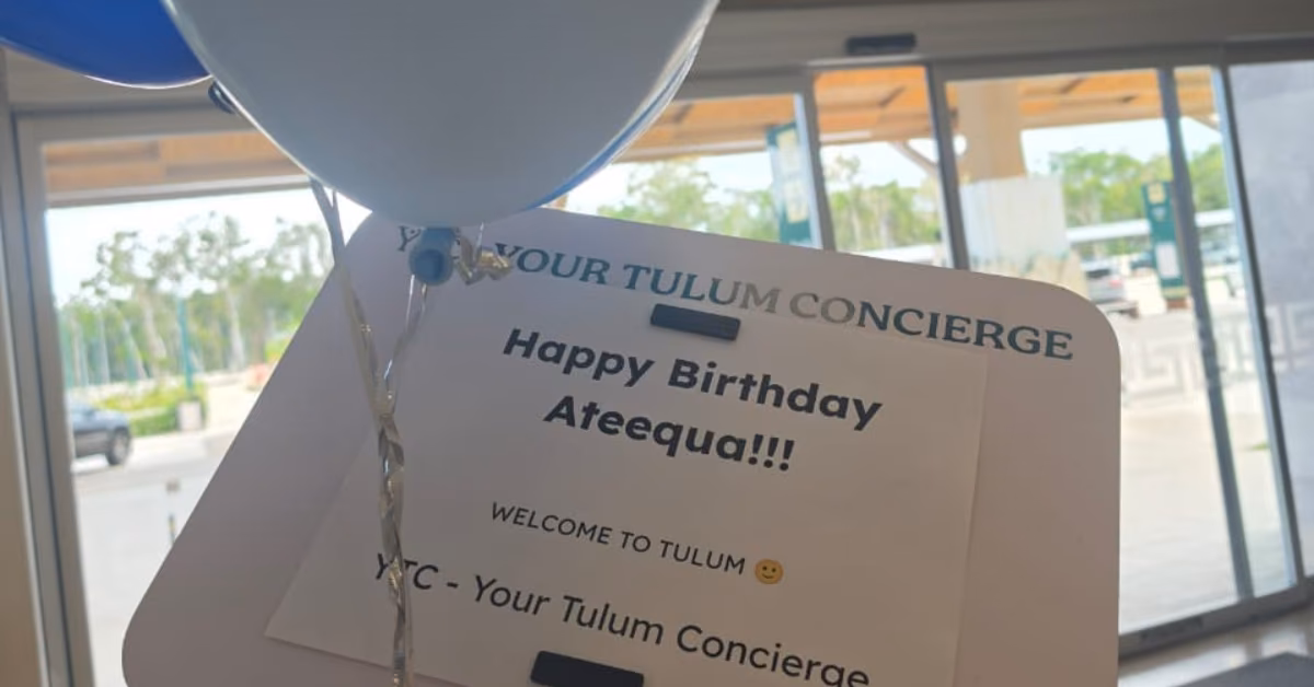Blue and white balloons with a “Happy Birthday Ateequa Welcome to Tulum” sign, outdoor view through glass doors behind.