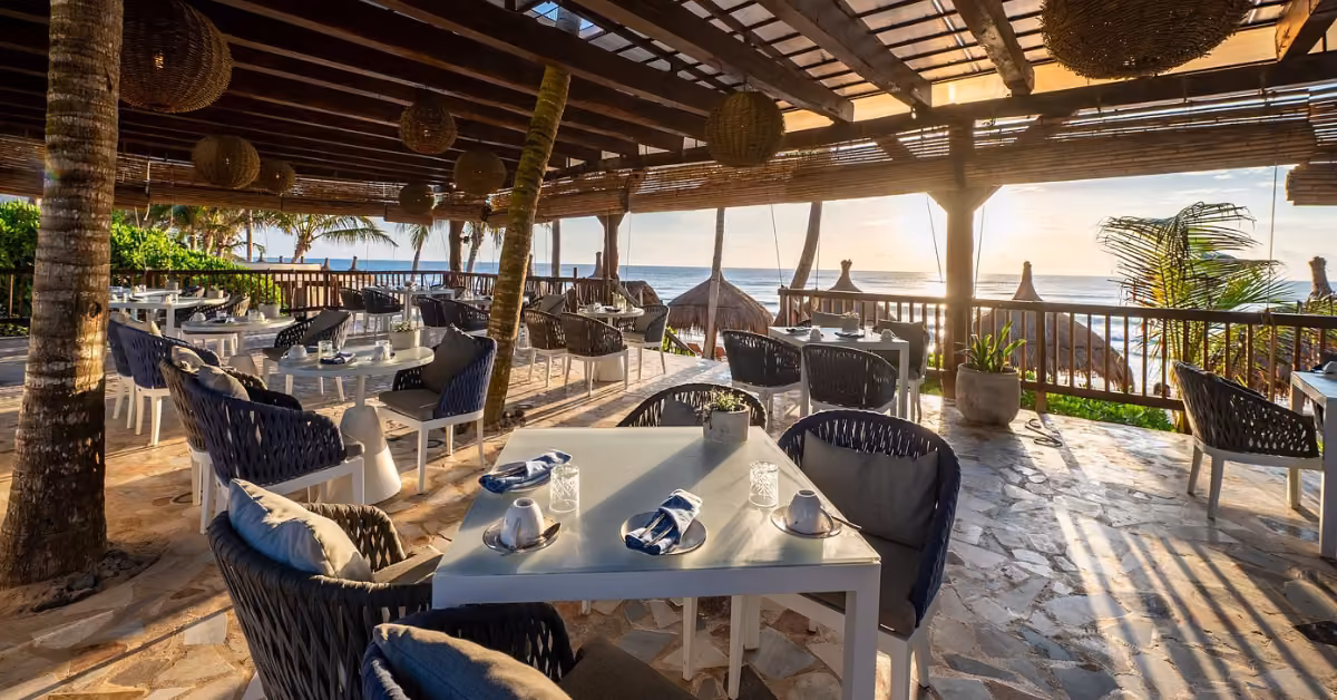 Beachside restaurant at sunset with white tables, gray chairs, and table settings under a wooden roof in a relaxed atmosphere.