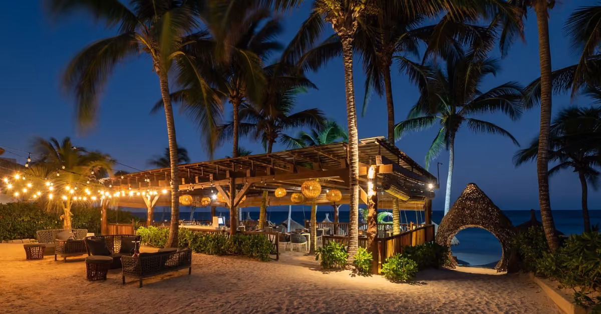 Beachside lounge at night with string lights, palm trees, wicker chairs, and a wooden bar. A cozy, illuminated hut and ocean waves are visible in the background, creating a relaxed, tropical atmosphere.