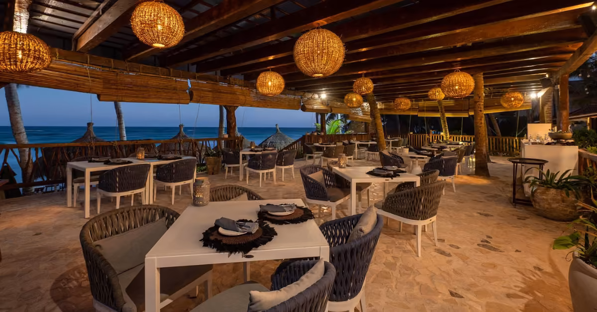 Beachfront restaurant at dusk with wicker chairs, white tables, pendant lights, wood beams, and an ocean view with palm trees.