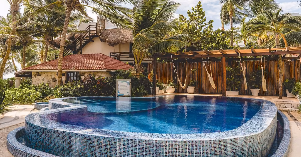 Resort pool with blue tiles at Ana y José Beach Club, surrounded by palm trees, hammocks, and a red-roofed building.