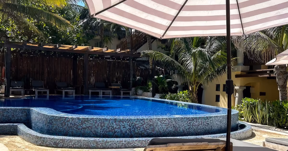 A curved pool at Ana y José Beach Club is ringed by lounge chairs, palm trees, and striped umbrellas under tropical sun.