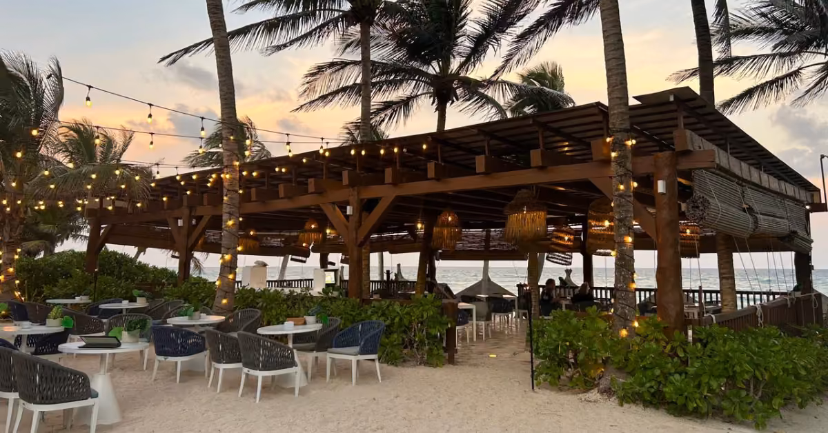 Beachside restaurant with wooden beams, string lights, palm trees, and outdoor seating on sand at sunset by the ocean.