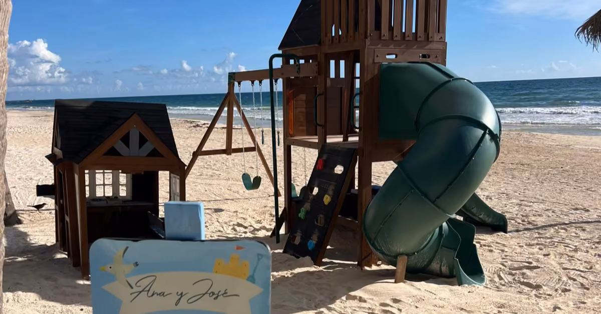 Wooden playground with slide, swings, and playhouse on a sandy beach by the ocean, “Ana y José” sign in front.