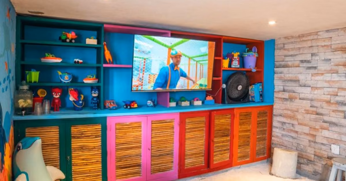 A bright playroom with colorful shelves, toys, blue walls, stone accents, a TV on, and bamboo-front cabinets exudes a playful vibe.