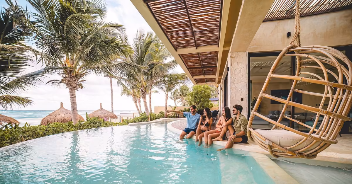 Four people relax by a pool at Ana y José Beach Club, with palm trees, huts, a wicker chair, and the ocean in view.