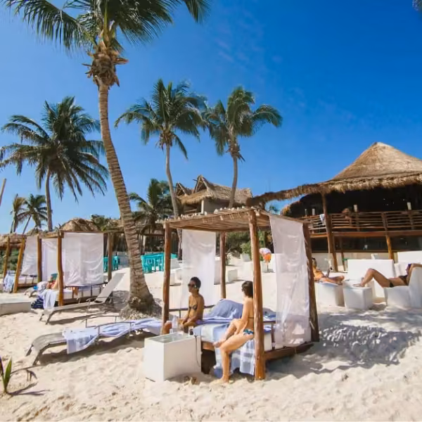 People lounge under cabanas on a palm-lined sandy beach near a thatched-roof beach club, all beneath a clear blue sky.
