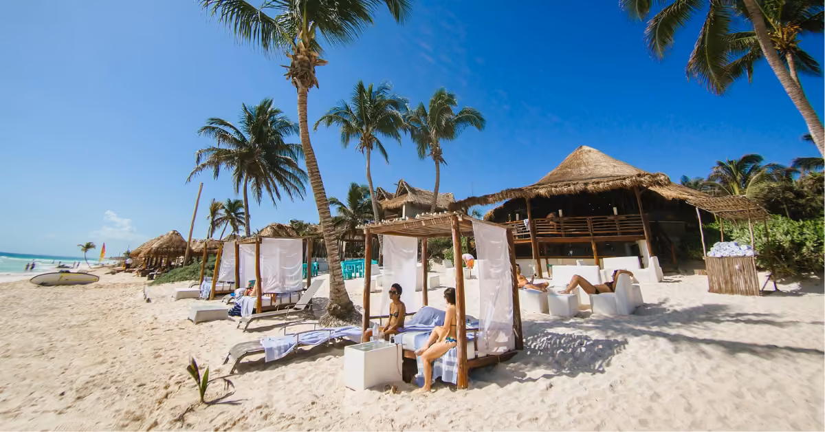 People lounge under cabanas on a palm-lined sandy beach near a thatched-roof beach club, all beneath a clear blue sky.
