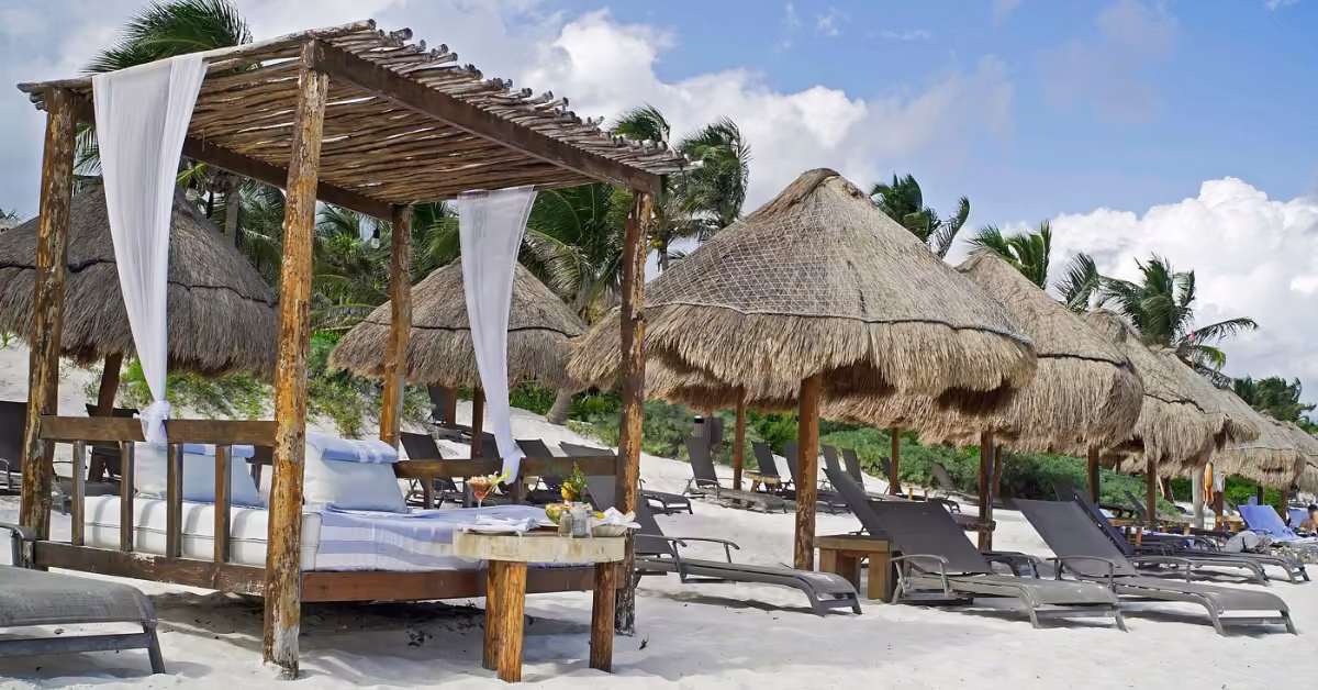 Cabanas with white curtains and thatched umbrellas line a sandy shore, with lounge chairs facing the sea under palm trees and blue sky.