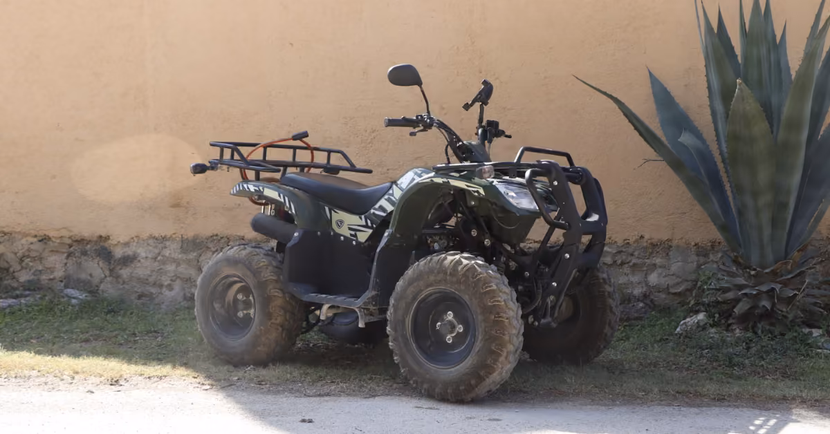 A green and black ATV with muddy tires sits on grass beside a beige wall and a large agave plant.