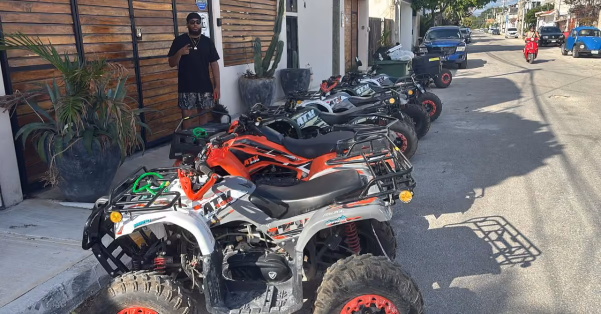 ATVs are lined up on a sunny street as a person in dark clothes stands by a wooden fence with large potted plants nearby.
