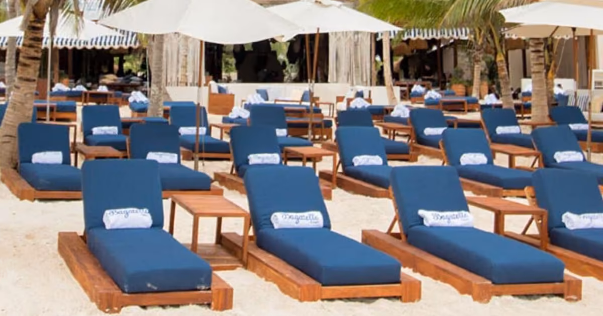 Rows of wooden lounge chairs with blue cushions and rolled towels are arranged on a sandy beach under white umbrellas at Bagatelle Beach Club, surrounded by palm trees and shaded structures in the background.