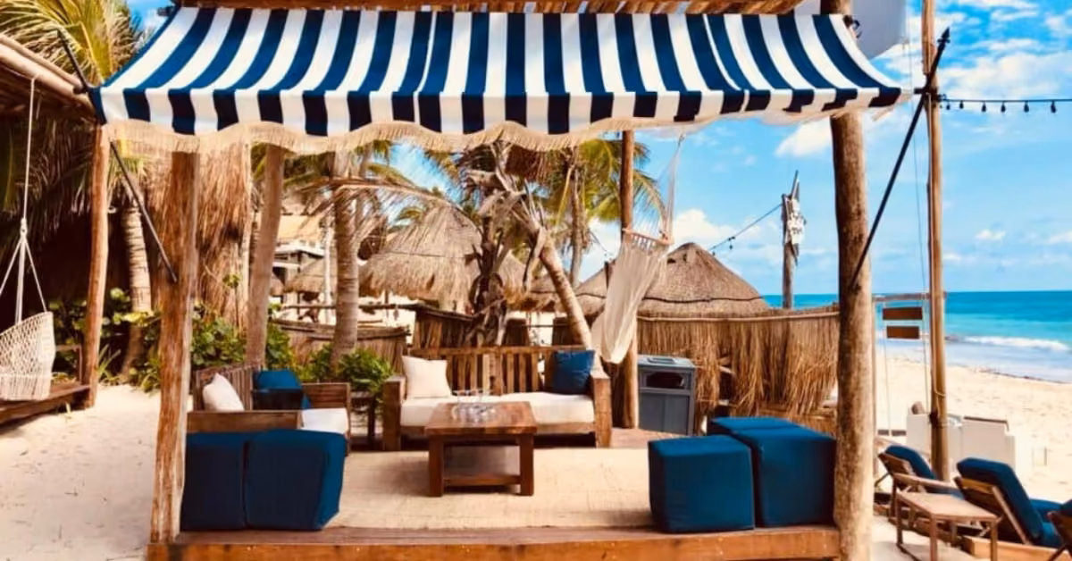A shaded beach cabana with blue and white striped canopy, wooden furniture, and blue cushions sits on sandy Bagatelle Beach Club near palm trees, huts, and lounge chairs by the ocean under a sunny sky.