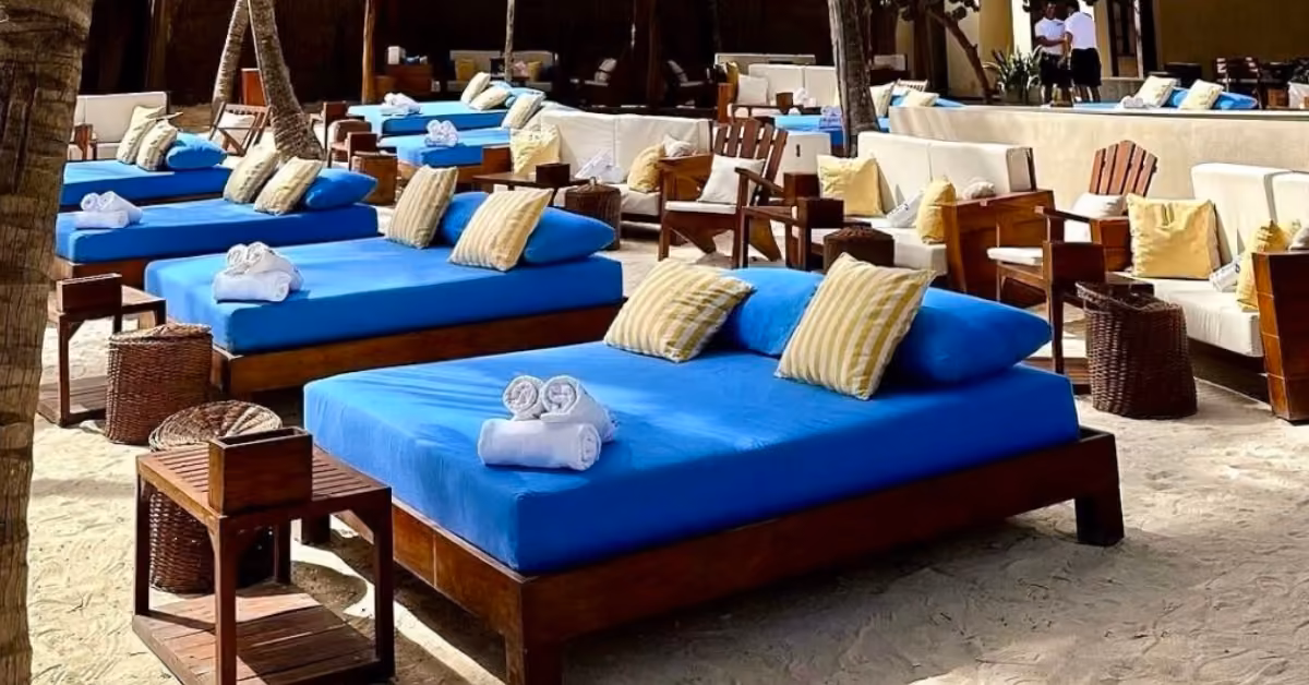 Outdoor lounge area on sandy ground at Bagatelle Beach Club, with rows of wooden beds covered in blue mattresses, yellow-striped pillows, and rolled white towels, surrounded by wooden chairs and wicker tables. People are visible in the background.