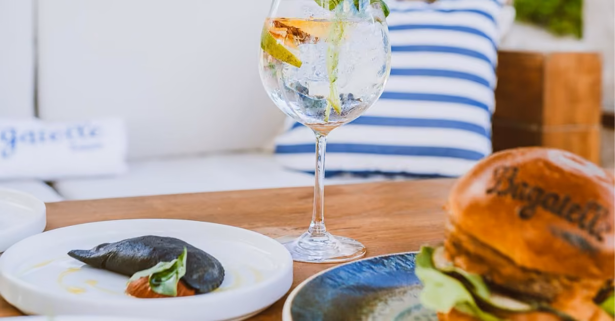 A close-up of a gourmet meal at Bagatelle Beach Club on a wooden table, featuring a burger, a black-colored dumpling, and a glass of gin and tonic with herbs and lemon, with striped pillows in the background.