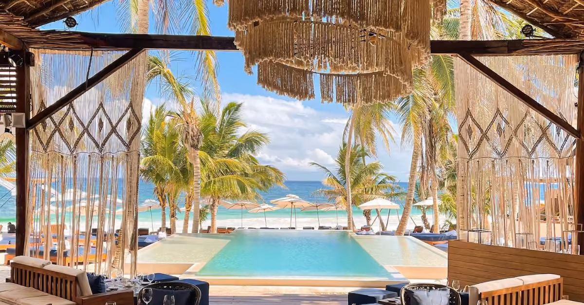 Open-air lounge with cushioned seating overlooks an infinity pool, palm trees, and a sandy beach with umbrellas at Bagatelle Beach Club. Decorative fringe hangs from the ceiling, framing a view of the blue ocean and sky.