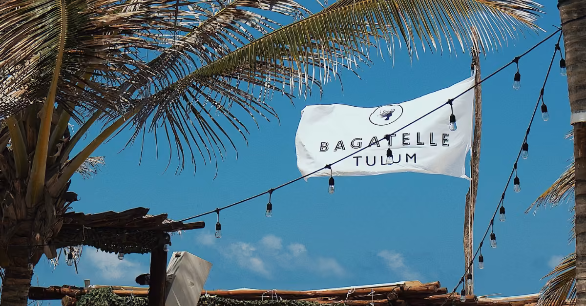 A white flag with the text Bagatelle Tulum, representing the iconic Bagatelle Beach Club, waves among palm trees under a blue sky, with string lights hanging in the foreground.
