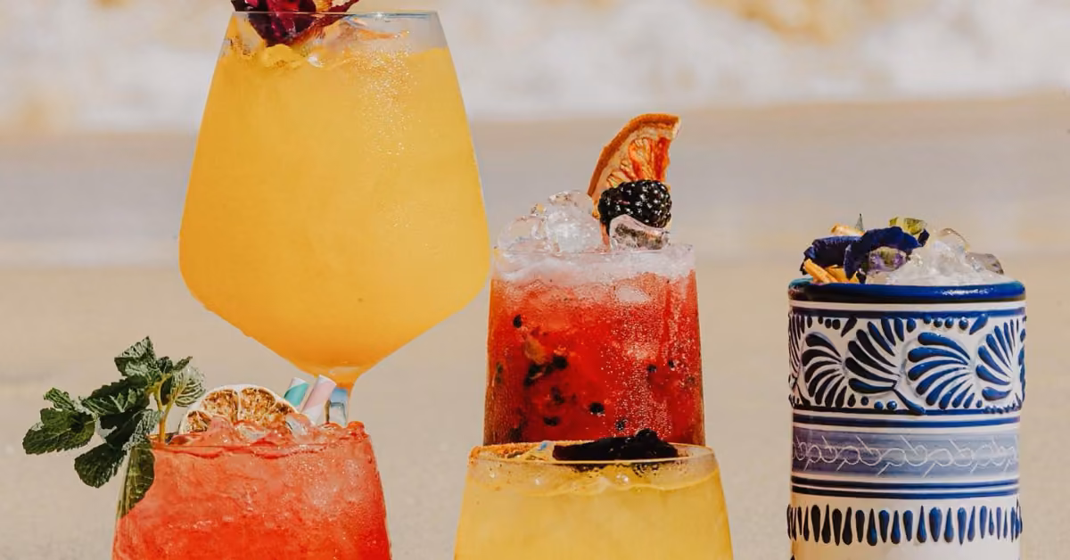 Four colorful cocktails in different glasses sit on a sandy surface at Bagatelle Beach Club, decorated with fruit, herbs, and flowers. One drink is in a blue patterned cup; the others are in clear glasses with ice and vibrant garnishes.