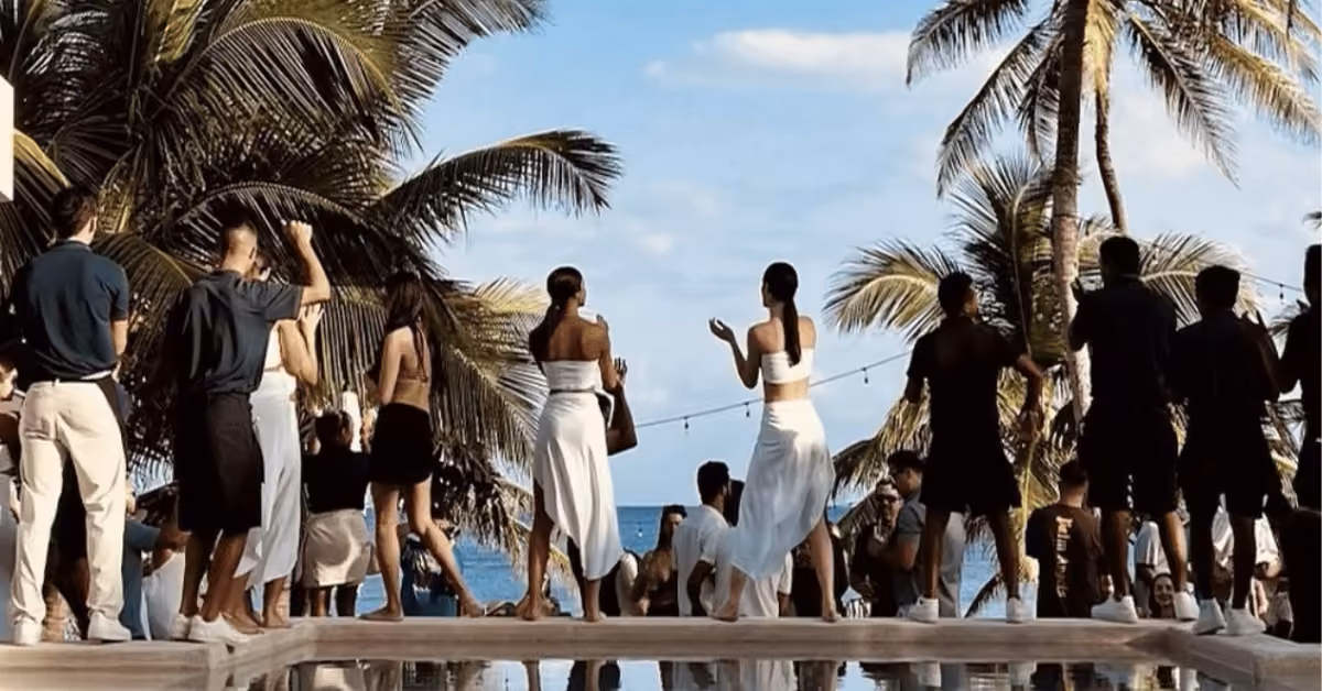 A group of people dressed in summer clothes gather by the poolside at Bagatelle Beach Club, dancing and socializing under palm trees with the ocean and blue sky in the background.