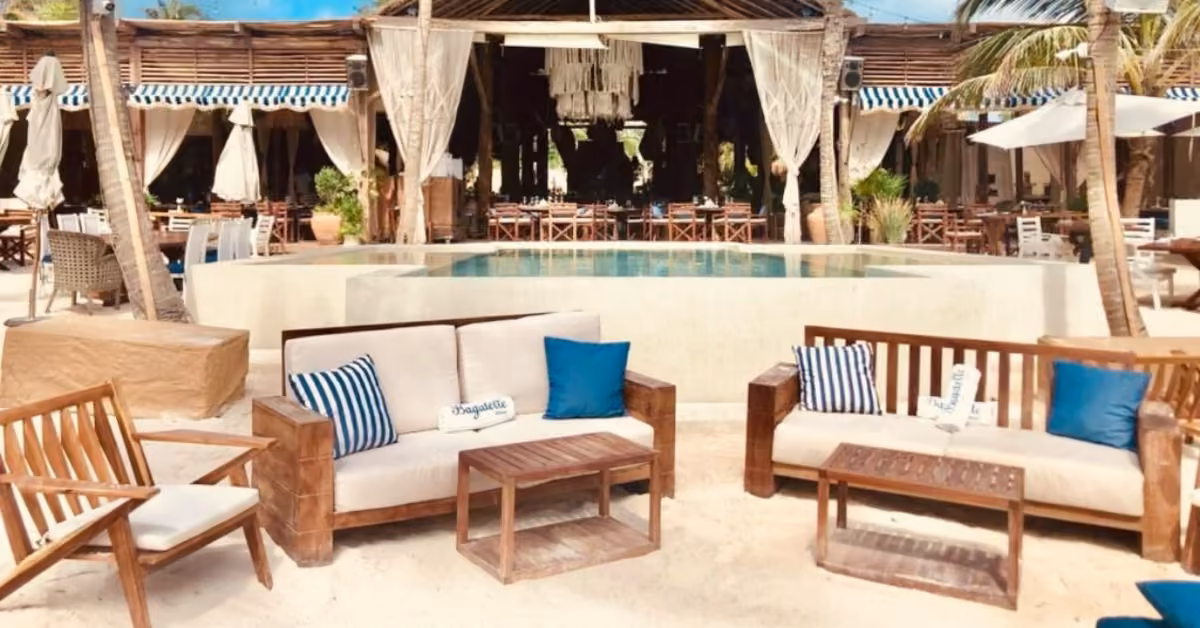 Outdoor beach lounge at Bagatelle Beach Club with wooden sofas, blue and white cushions, and small tables set on sand in front of a stylish open-air restaurant with a pool and palm trees. Relaxed, tropical atmosphere.