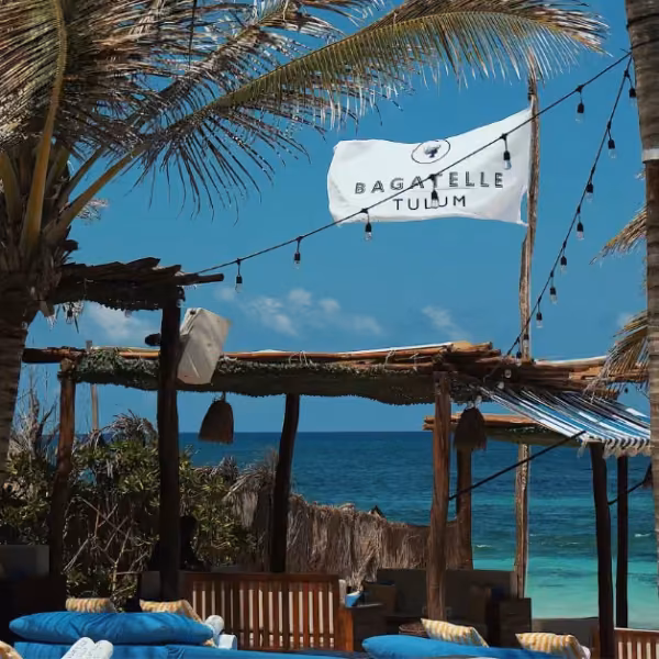 A beachside lounge area with wooden seating, blue cushions, and hanging lights shaded by palm trees. A white flag reading Bagatelle Tulum flies above, offering classic beach clubs vibes with turquoise ocean and clear sky in the background.