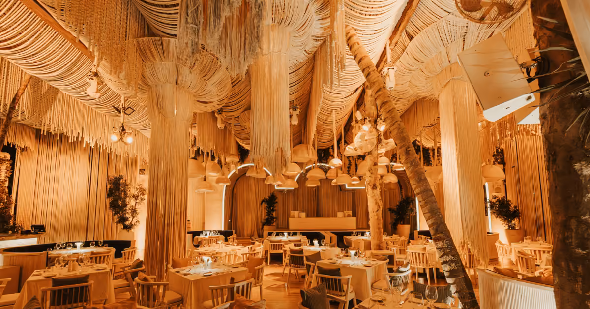 A restaurant interior with warm lighting, wooden tables and chairs, and elaborate ceiling decorations made of hanging ropes, much like the cozy and artistic atmosphere found at Bagatelle restaurant.
