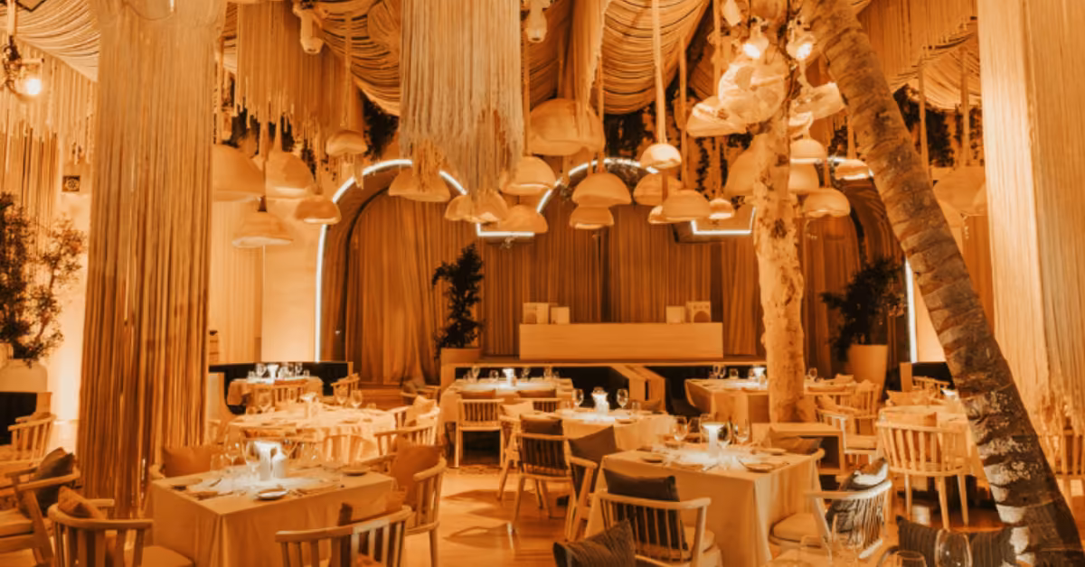 A warmly lit bagatelle restaurant with wooden chairs and tables, draped fabric and hanging lamps from the ceiling, and natural elements like tree trunks and plants creating a cozy, elegant atmosphere.