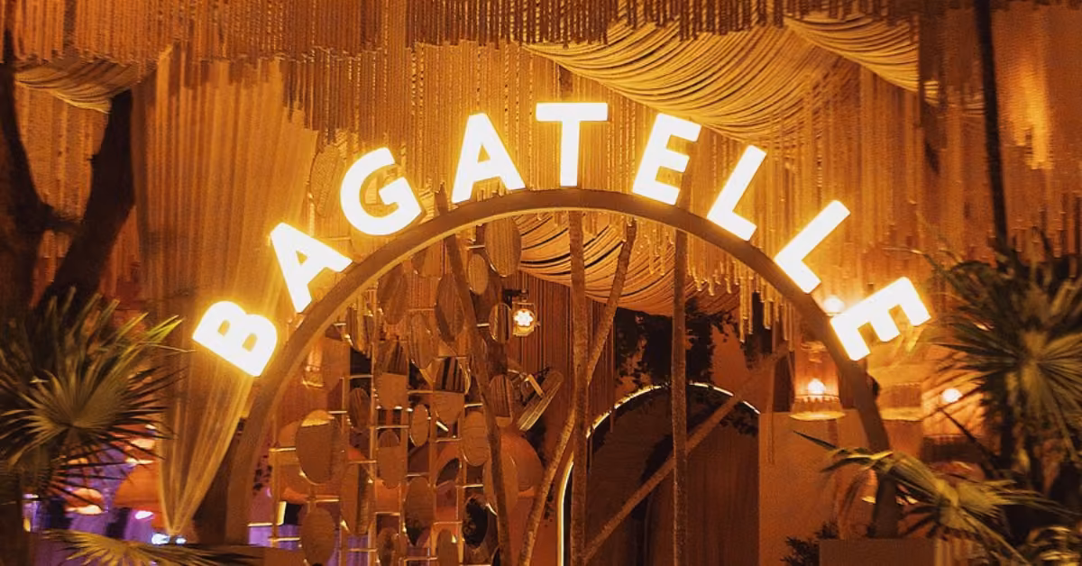 A glowing sign reading BAGATELLE arches over the bagatelle restaurant entrance, decorated with hanging lights, draped fabric, and lush plants, creating a warm and inviting atmosphere at night.