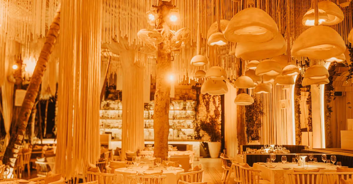 A warm, ambient Bagatelle restaurant interior with hanging mushroom-shaped lights, draped fabric, a central tree, white tables and chairs, and a well-lit bar area in the background.
