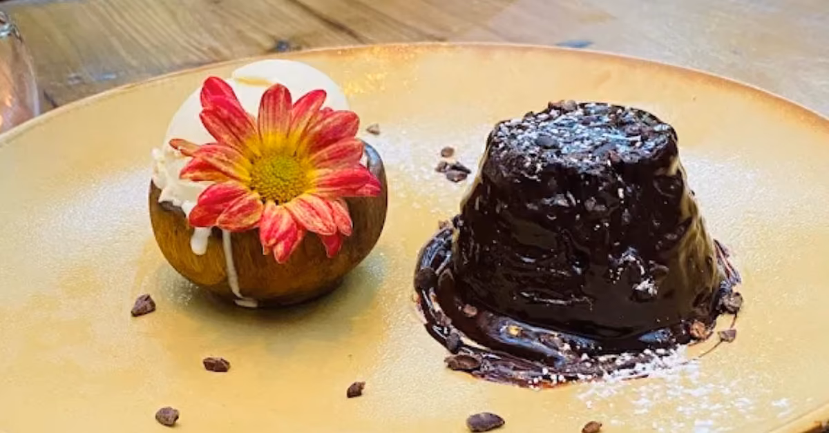 Vanilla ice cream with a pink and yellow flower sits beside glossy chocolate lava cake on a tan plate with cocoa nibs.