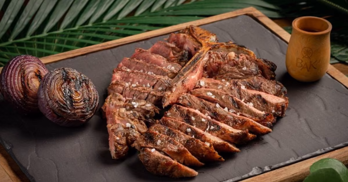 Grilled steak slices on a slate with coarse salt, roasted red onion halves, and a wooden cup on a tray with green leaves.