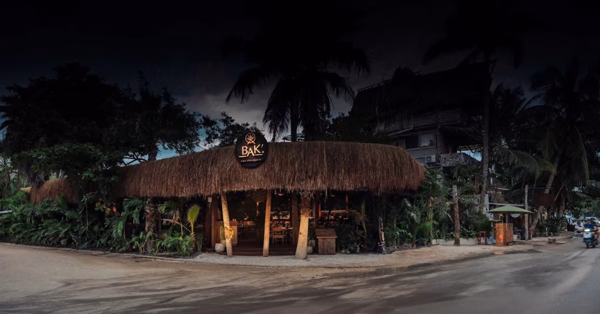 A warmly lit rustic restaurant with a thatched roof and tropical plants, “BAK” sign out front, palm trees against the night sky.