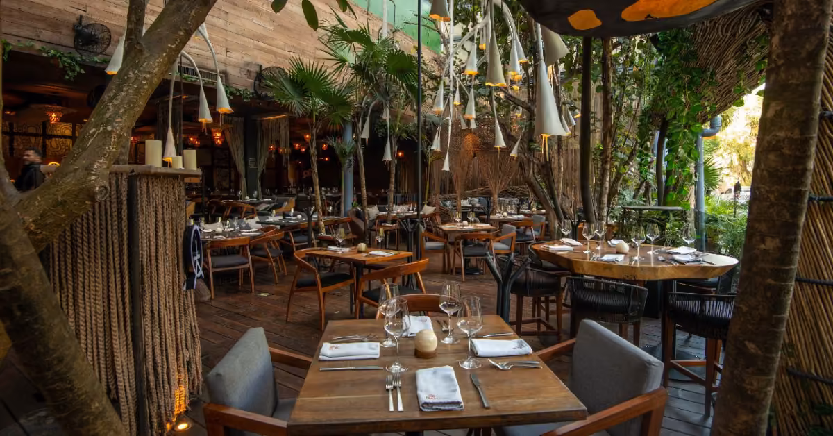 Bak restaurant’s outdoor seating features wooden tables, greenery, and warm lights for a cozy, inviting dining experience.