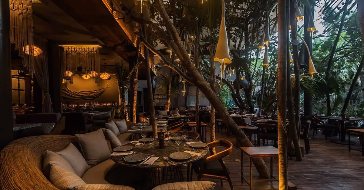 A dimly lit restaurant with cozy seating, wooden tables, hanging lights, and lush greenery creates a warm, inviting vibe.