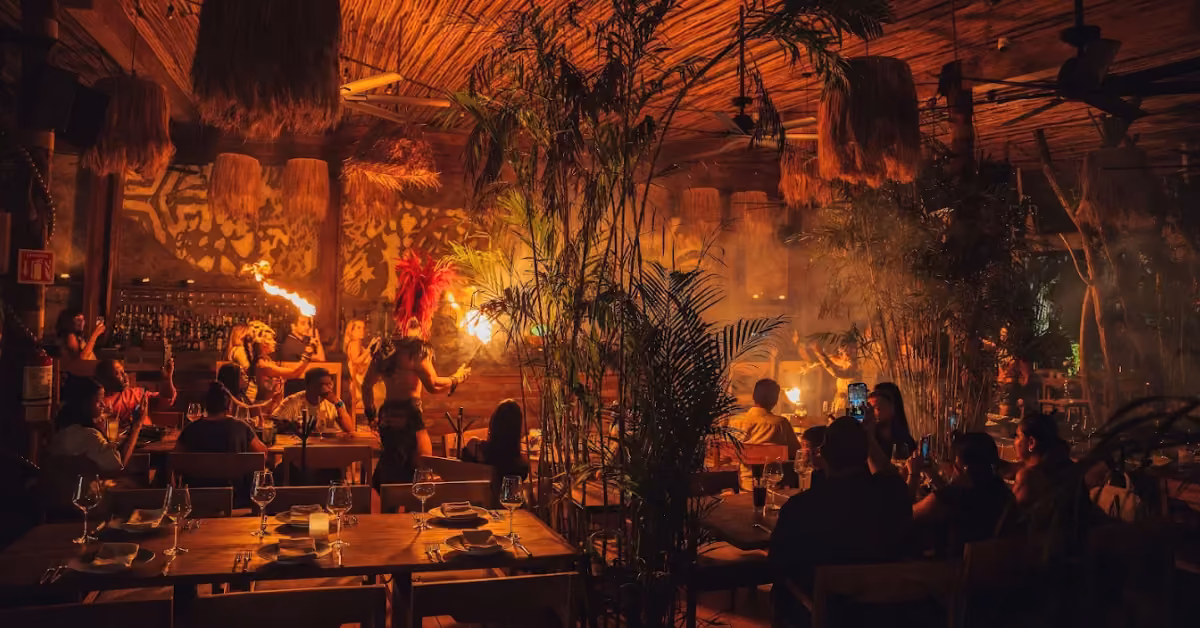 Cozy restaurant with wood decor, diners at tables, lush plants, and performers in colorful costumes entertaining with fire.