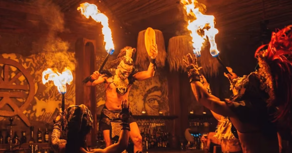 Four dancers in elaborate costumes and headpieces perform indoors with flaming torches, warm light, mural, and bar behind them.