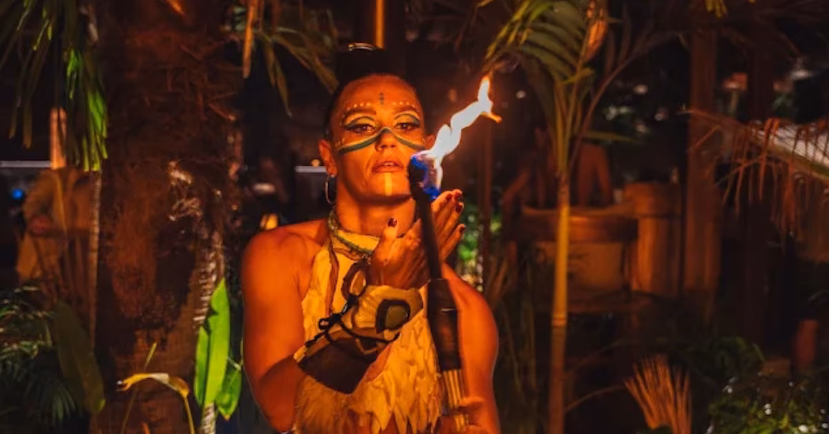 A performer with face paint and tribal attire holds a flaming torch near their mouth, surrounded by lush plants in dim lighting.