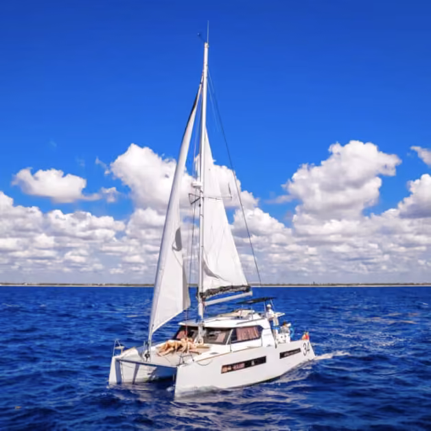 A white sailboat with raised sails glides on deep blue water under a bright sky as someone relaxes on the front deck.