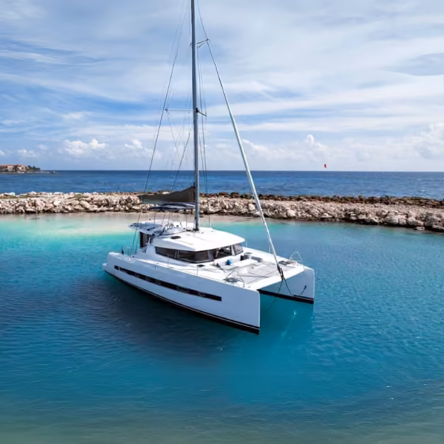 A white catamaran floats on calm turquoise water near rocky shore, beneath a partly cloudy sky and tranquil ocean backdrop.