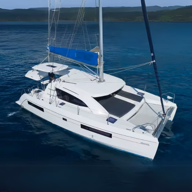 A white catamaran with blue accents floats on clear, calm water near a lush green coast under a bright, cloudless sky.