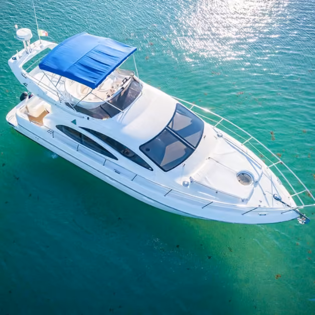 A white yacht with a blue canopy floats on clear turquoise water, viewed from above in bright sunlight.