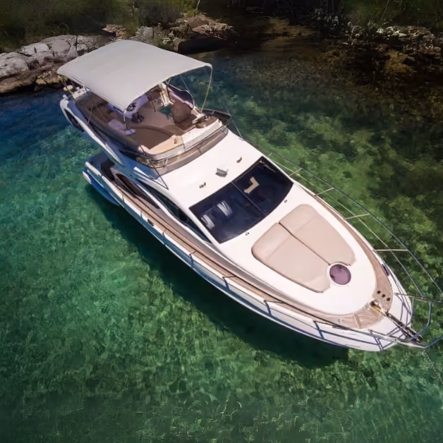 A white motor yacht with a canopy anchors in clear turquoise water near a rocky shore, surrounded by lush greenery.
