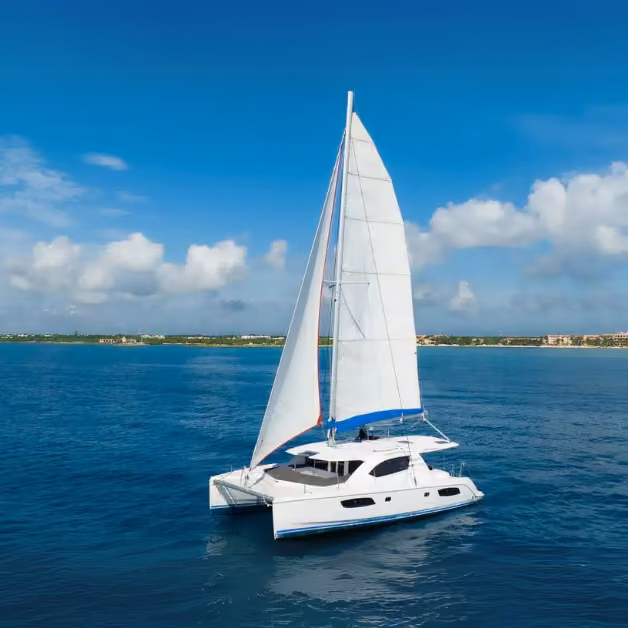 A white catamaran with blue accents sails on calm ocean water under a clear sky, heading toward the distant shoreline.
