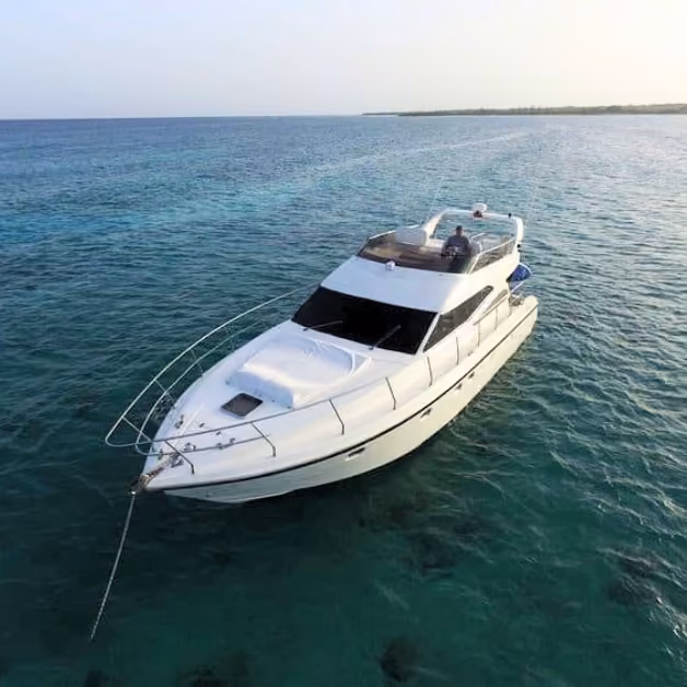 A white yacht anchored in turquoise water near the coast. One person relaxes on the upper deck under a clear, calm sky.