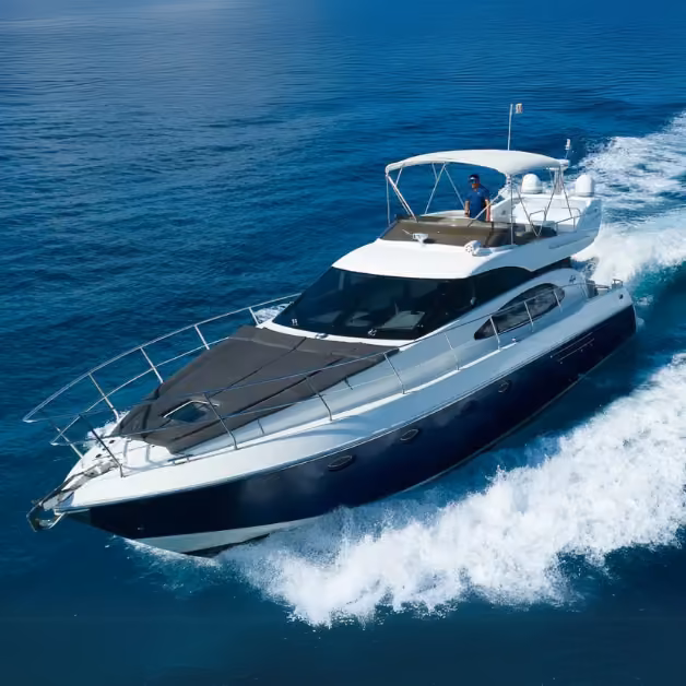 A sleek yacht glides over blue water, white waves trailing behind as someone steers from the sunny upper deck.