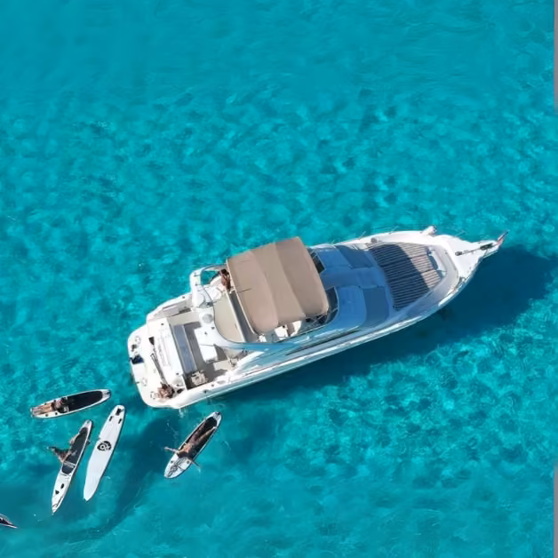 A white yacht on turquoise water is surrounded by people paddling boards and kayaks on a sunny day, seen from above.